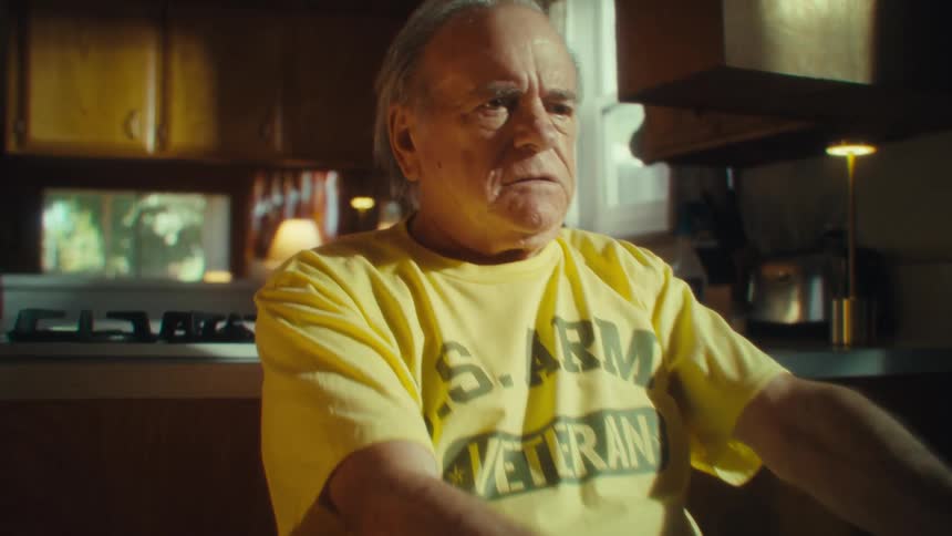 An elderly man sits indoors in the kitchen dining area sorting through pieces of paper at midday