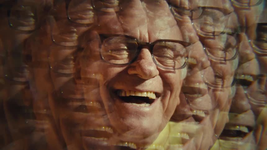 An elderly man wearing glasses and a yellow shirt scrolls through his phone indoors and suddenly bursts out laughing