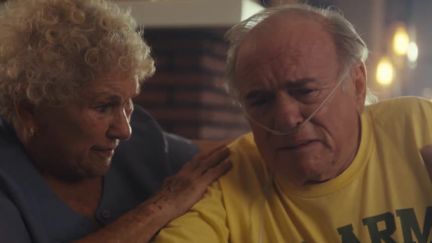 The video shows an elderly man with an oxygen cannula in his nose sitting at a table he is wearing a yellow tshirt and appears to be in distress as an elderly woman comforts him by placing her hand on his shoulder