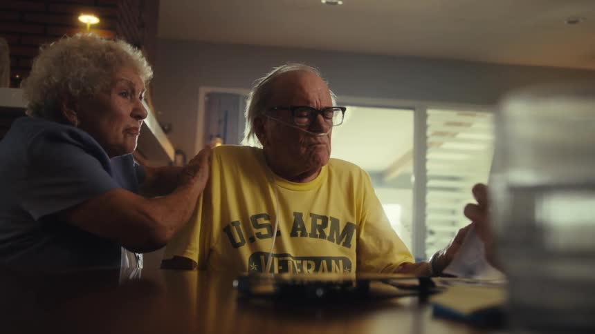 The video shows an elderly couple and a man sitting at a table the man is showing them some papers and the elderly man who is wearing a us army veteran tshirt and has an oxygen cannula under his nose is talking with his hands