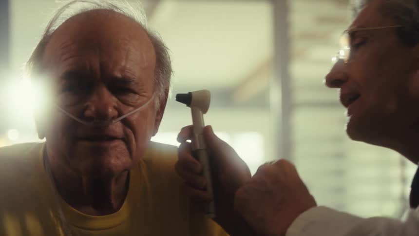 A doctor wearing glasses and a stethoscope examines the ear of a man who is wearing a yellow shirt and has a nasal cannula in his nose