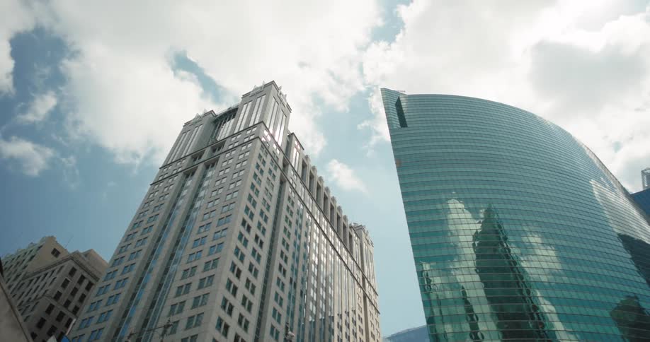 Two tall buildings against a partly cloudy sky with the reflection of the clouds and sky visible on the glass exterior of the second building