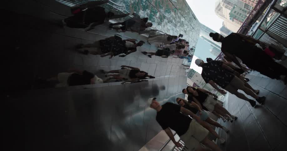 A group of people some walking and others standing are on a pedestrian bridge as the roof reflects the ocean view with the city skyline in the background at midday
