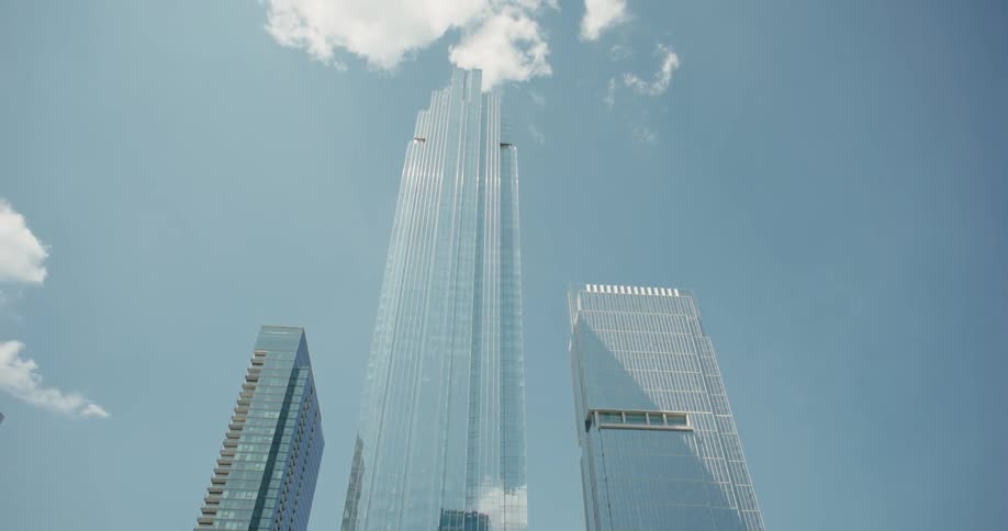 A camera travels underneath a dark metal bridge then emerges to show a close view of several tall modern glass buildings with a bright pale blue sky and scattered clouds in the background