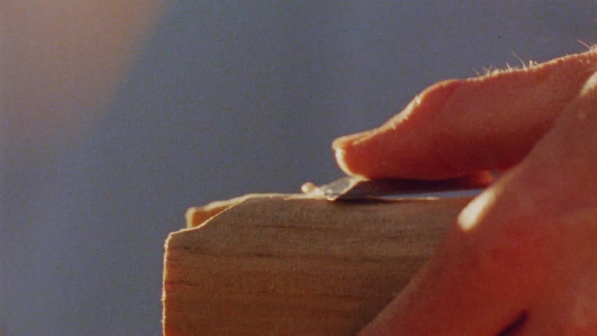 A persons hand guides a sharp tool along the edge of a wooden plank shaving off thin curls