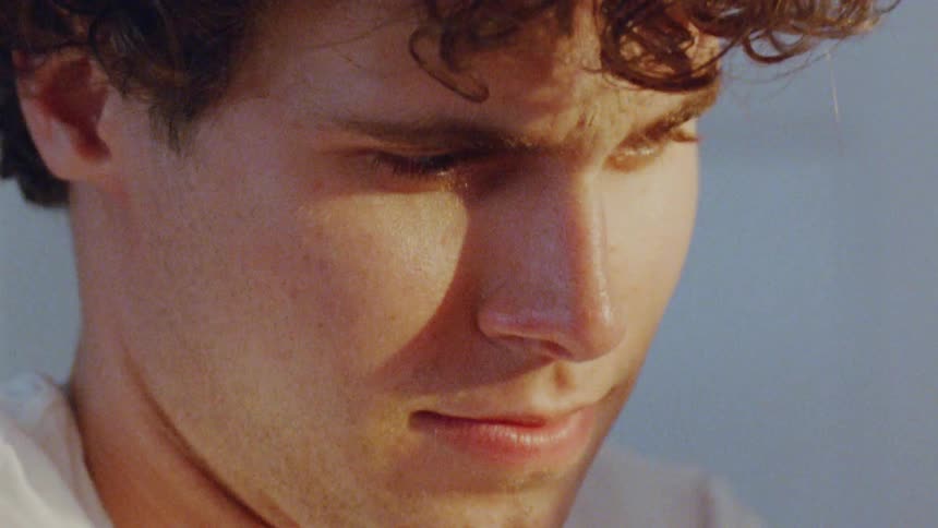 A person is seen in a closeup shot with their eyes looking down away from the camera the video captures a contemplative or somber mood the person has curly hair and the sunlight illuminates a portion of their face casting a warm glow