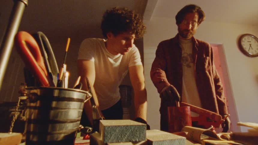 Two artists stand at a workbench in a bright room with the man on the right stirring a can of red paint with a paintbrush while the man on the left watches him