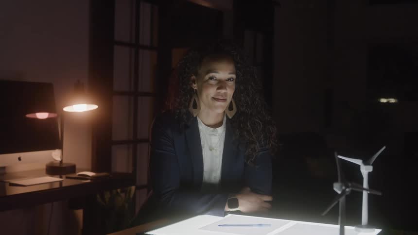 A smiling woman with long curly hair is sitting at a desk in a dimly lit office she is leaning on her arms which are crossed on top of an illuminated surface a small model of a wind turbine is visible to the right