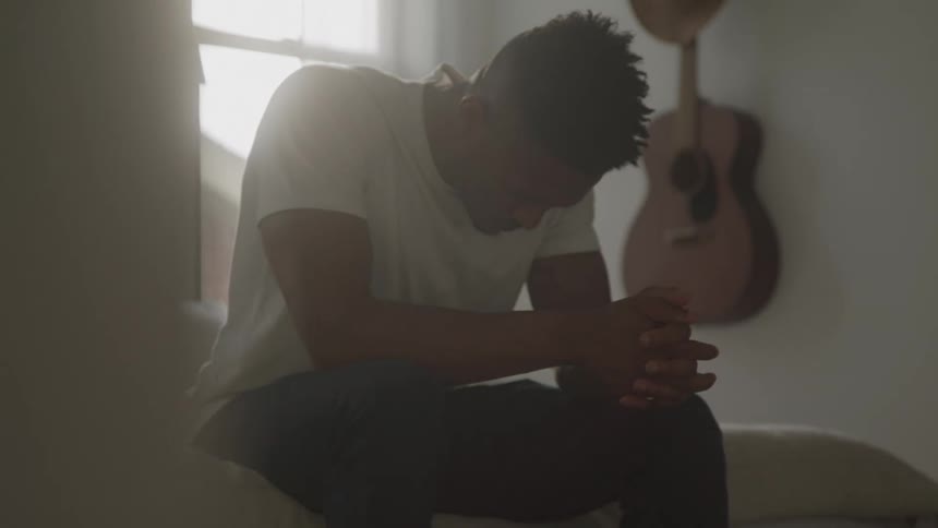 Man sitting on the edge of the bed with head down and hands clasped