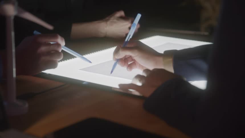 Hands point pens at a light table during a collaborative activity