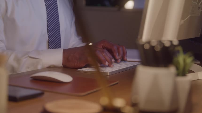 A close view of a person with dark skin shows them typing on a white keyboard with both hands with a white and blue striped tie and a white shirt visible