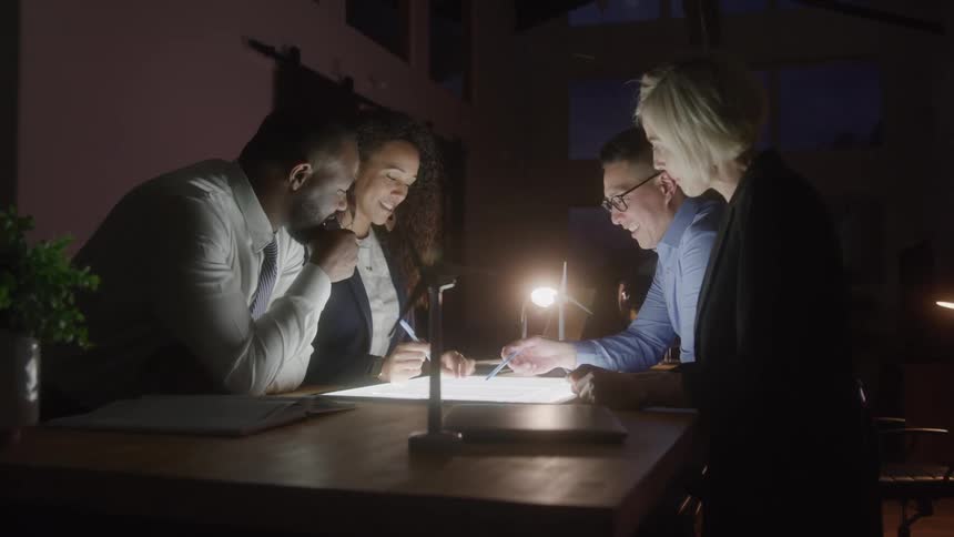 A diverse group of four coworkers two men and two women huddle together at a table in a dimly lit office looking down at a light box and a paper while talking and gesturing