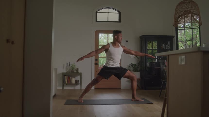 A man practices yoga in his living room holding a steady warrior pose