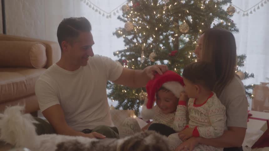 Family gathered in front of the christmas tree with children in festive pajamas and one wearing a santa hat