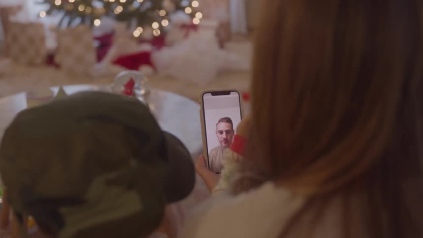 A mother and her son sit at the dining table as the mother holds a smartphone during a video call with the husband both wave at him