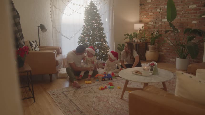 A young family interact playfully while sitting on the floor in front of a christmas tree in their living room during daytime
