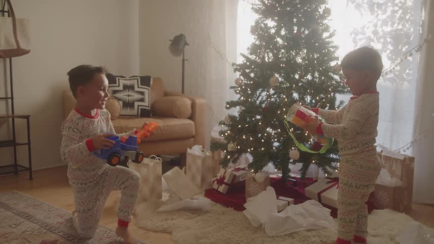 Two young boys in festive pajamas excitedly play with new toys beside a decorated christmas tree