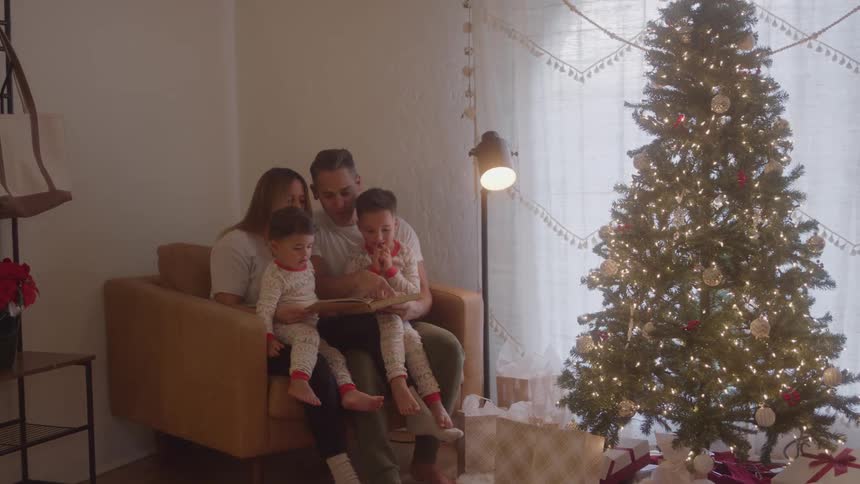 A family of four a couple and their two boys sitting together on a small couch near a christmas tree and a window as the father reads the children a story from a book