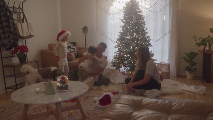 A family gathered in the living room the father and mother sitting two children standing in pajamas and a dog with one of the boys hugging the father