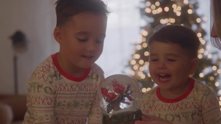 Close up of a young mother shaking a snow globe in front of her sons as they sit together in a living room during daytime