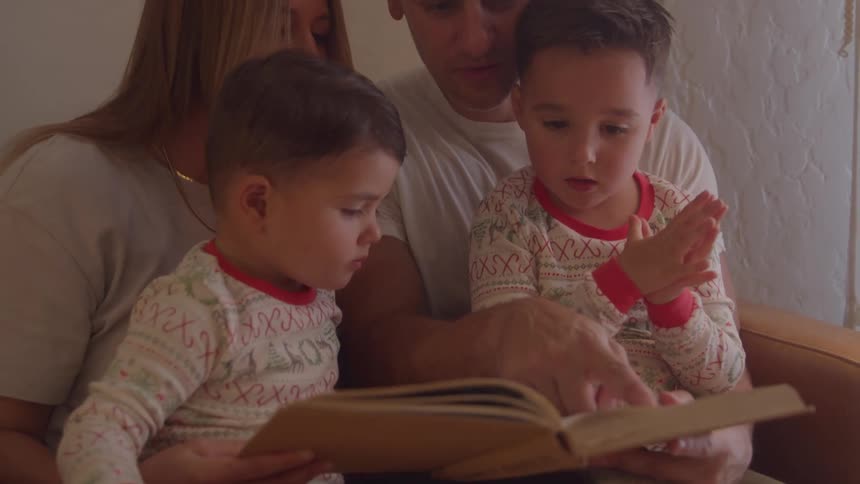 A couple sits on a couch at midday each holding one of their twin children on their lap as they read a book together