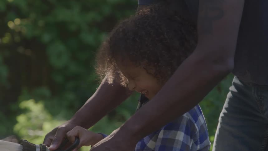 A person with a tattoo on their arm helps a child with curly hair and a plaid shirt spray a garden hose as the water sprays onto their hands and face