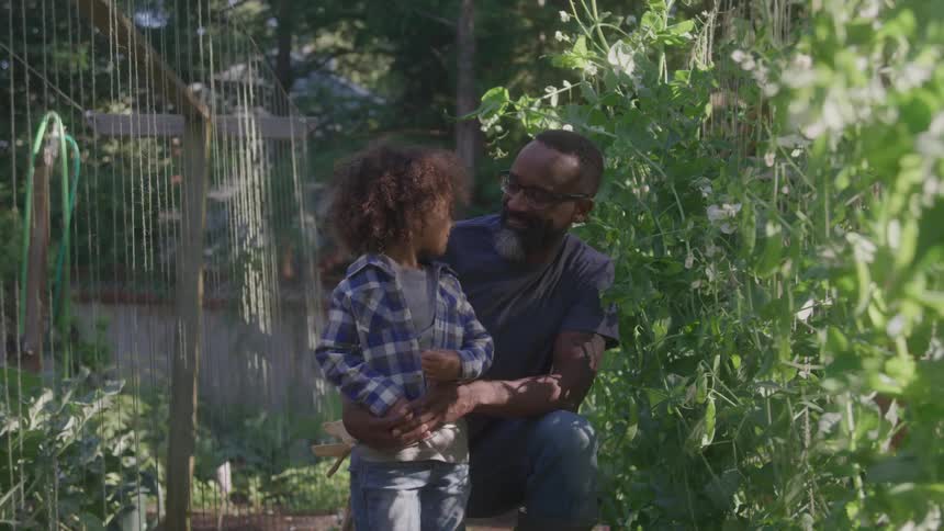 A man with dark skin wearing glasses and a blue shirt kneels in a garden next to a young child with long curly hair wearing a plaid shirt as they both look at each other and then smile at the camera