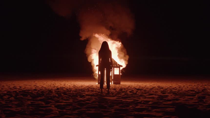 A woman stands watching a playhouse burn in flames in an open field at night