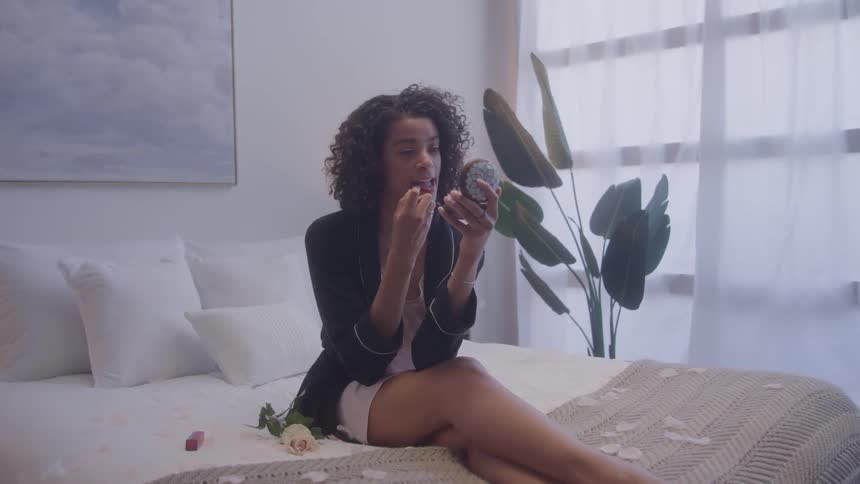 A woman sits on bed applying makeup in mirror