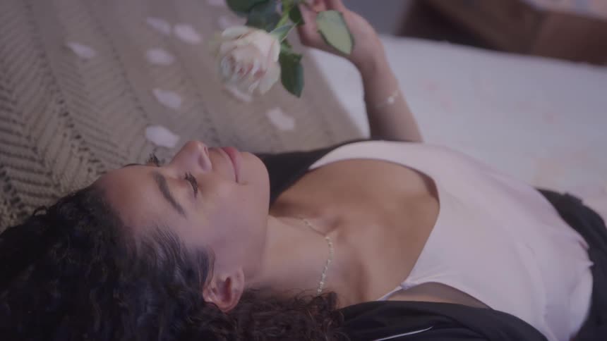 A woman lies on a bed sniffing a rose in the bedroom