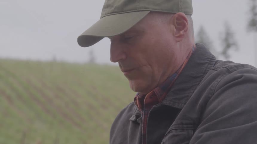 A man in a grass field wearing a cap doing work with his hands