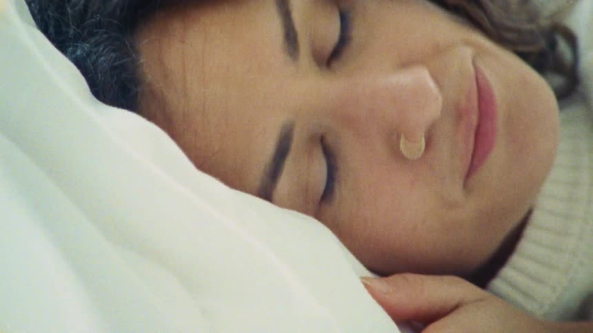 The video shows a closeup shot of a sleeping woman she has dark curly hair and a nose ring and a soft smile is visible on her face she appears to be peacefully asleep on a white pillow