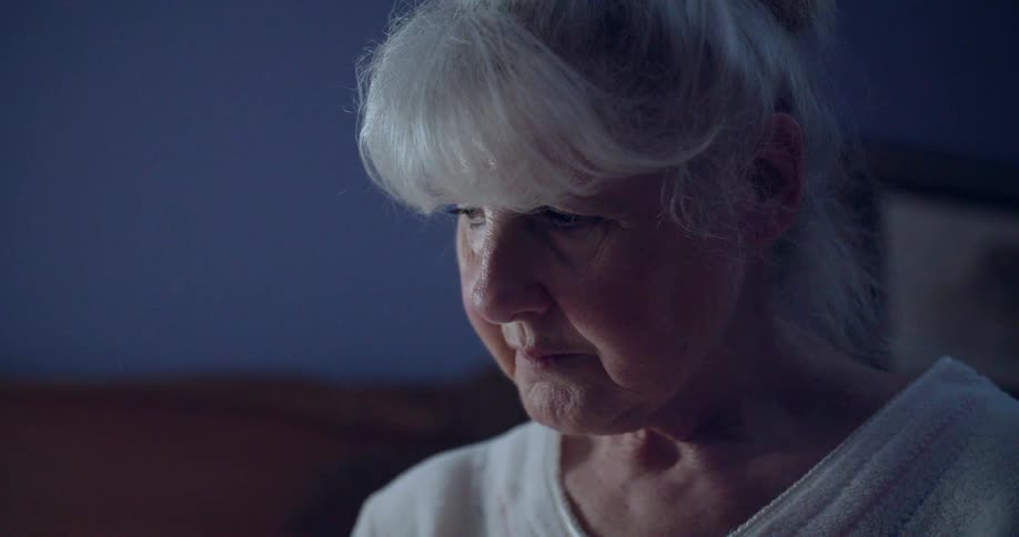 Elderly woman with white hair looks downward with a serious expression