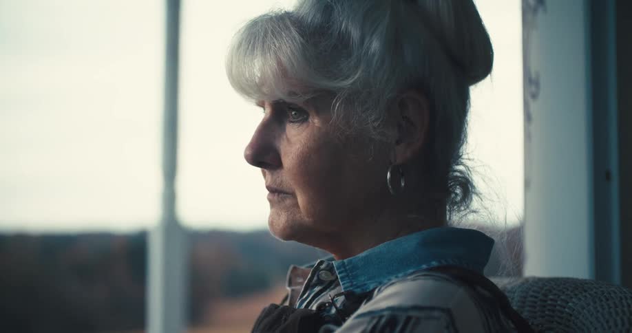 A side profile of an older woman with whiteblonde hair styled in a bun is shown she is looking out a window and the light from outside illuminates her face she appears to be looking out a window
