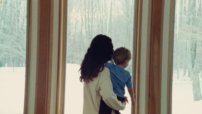 Woman holds a child in her arms while looking at the snowy landscape through the window