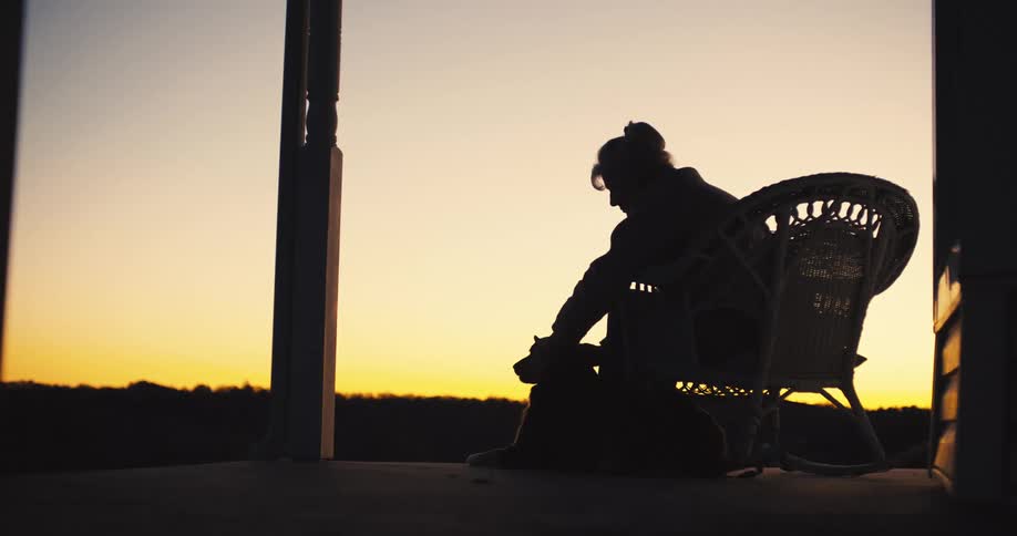 The woman pets her dog on the porch as the sun rises
