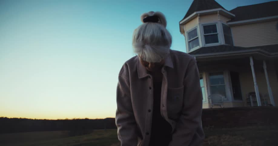 Elderly woman smiling and waving in front of a house at sunset