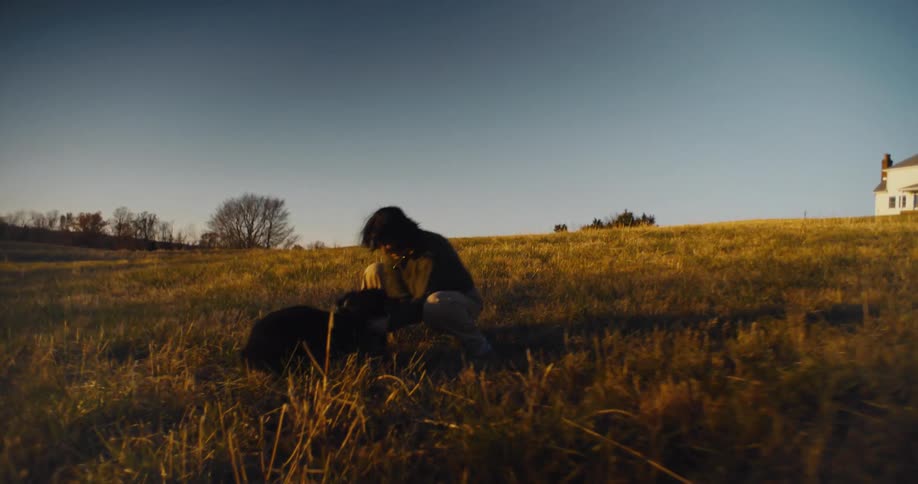 A woman wearing a brown sweater and light pants is crouched down in a field of dry grass playing with a black dog while a distant white house is visible on a hillside behind them