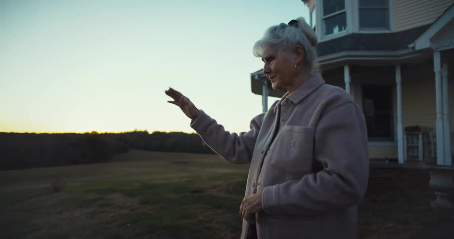 An elderly woman standing in front of her house waving to someone