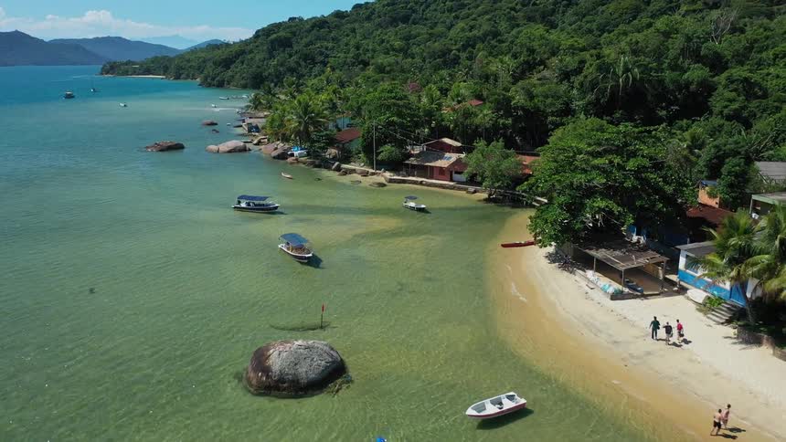 An aerial shot captures a scenic bay with clear turquoise water the video shows a sandy beach with several small boats floating nearby the beach is bordered by a lush green forest on one side and the calm ocean on the other