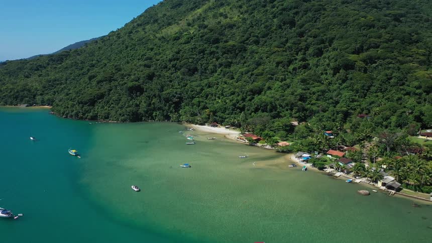 Boats float in the coastal water near a town located at the foot of a forested mountain on a sunny summer day
