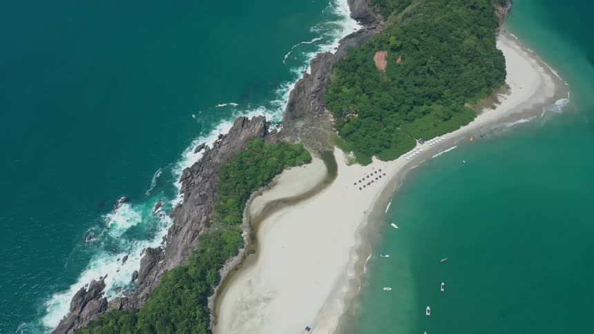 A high aerial view shows a long stretch of land jutting out into a turquoise colored ocean with a green and rocky shoreline a small white sand beach with umbrellas and chairs and several boats floating nearby