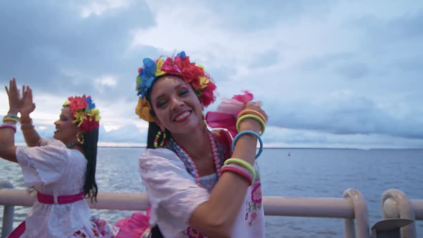 A female performer wearing a white dress a bright floral headdress and colorful beaded necklaces smiles at the camera as she dances with other performers next to a body of water in the evening