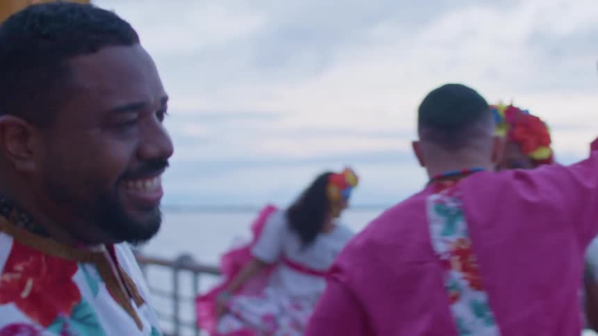A group of people of different races all wearing the same attire dance excitedly at the bay area while another group plays various musical instruments at midday