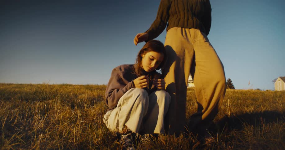 Girl sitting on the grass leans against the legs of another standing person