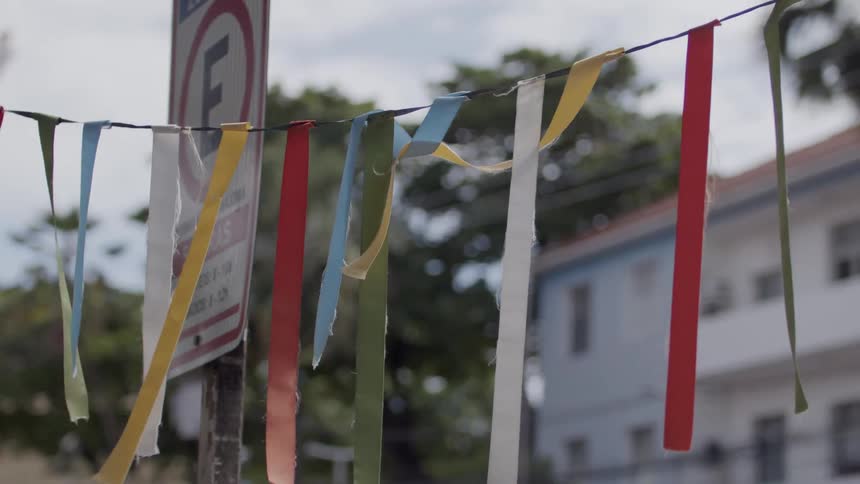A strand of multicolored fabric ribbons hangs on a wire between two poles fluttering in the wind with a blurred white church steeple and buildings visible in the background
