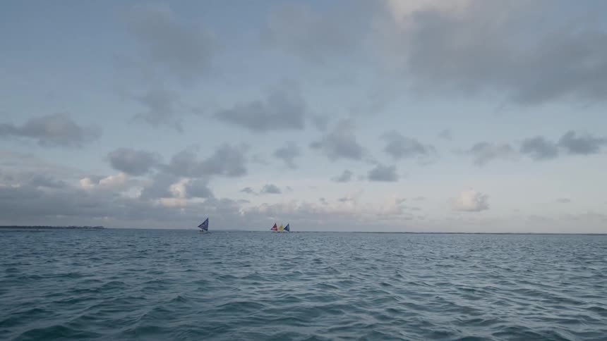 A low aerial shot flies over the open ocean towards two small sailboats with full sails visible on the distant horizon under a cloudy sky