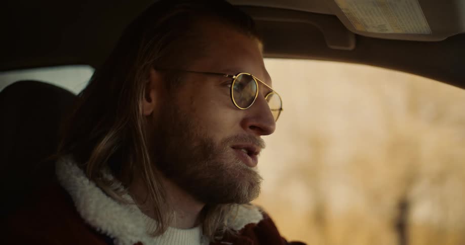 A man with long blond hair and a beard wearing a brown jacket with a white fleece collar and gold rimmed glasses sits in the drivers seat of a car turning his head to the side and smiling as he speaks