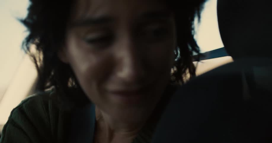 A young woman sits in the passenger seat of a moving car with her seat belt on leaning back with tears in her eyes as she cries at midday