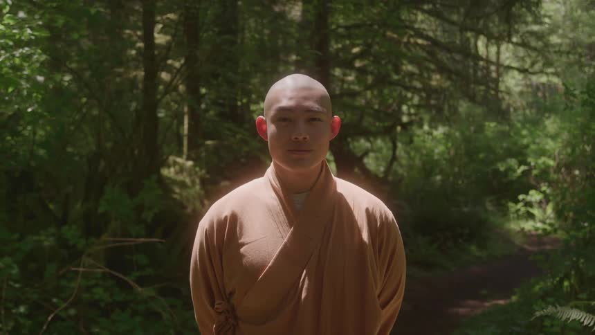 A monk wearing robes stands still on a dirt path in a sundappled forest and looks directly into the camera
